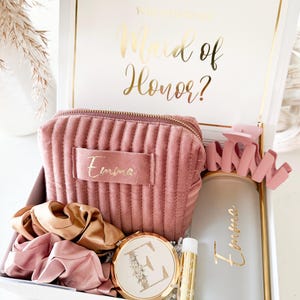 May include: A white gift box with gold trim and the text "Will you be my Maid of Honor?" in gold script. Inside are a pink velvet cosmetic bag, scrunchies, a compact mirror, a lip balm, and a frosted glass tumbler, all personalized with the name "Emma".