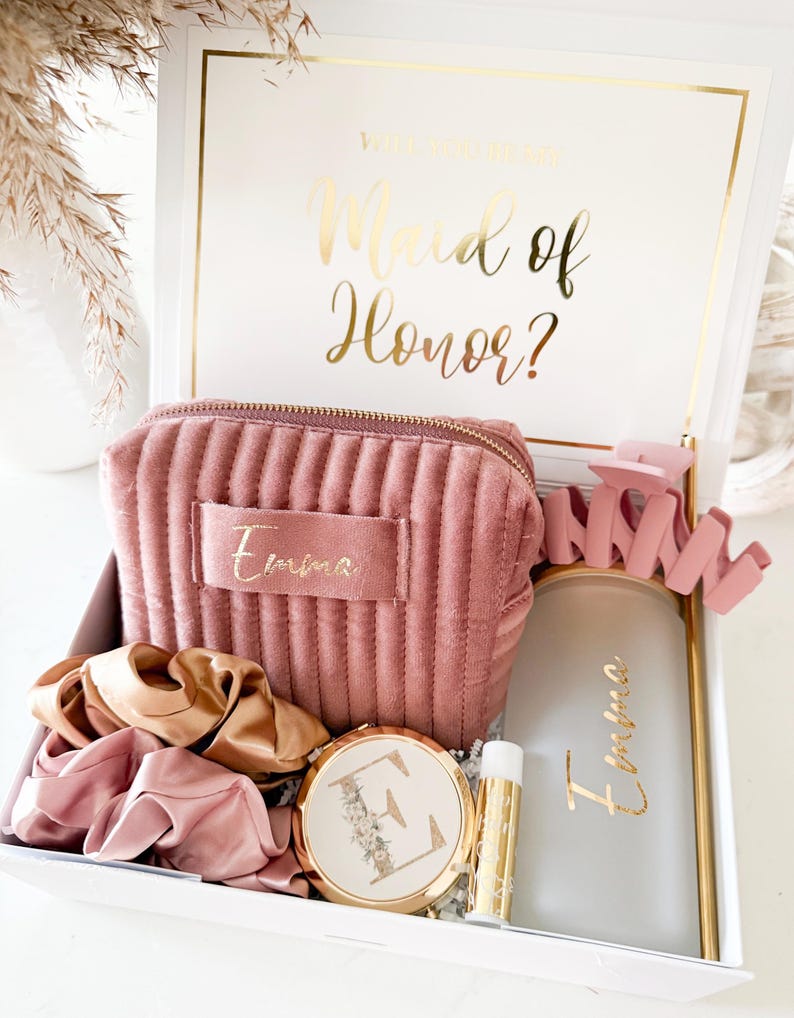 May include: A white gift box with gold trim and the text "Will you be my Maid of Honor?" in gold script. Inside are a pink velvet cosmetic bag, scrunchies, a compact mirror, a lip balm, and a frosted glass tumbler, all personalized with the name "Emma".