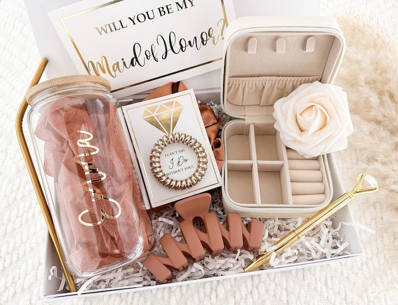 May include: A white gift box filled with items for a maid of honor. The box includes a glass water bottle with a gold lid and the name "Emma" written on it, a small jewelry box with a rose inside, a card that says "I can't say I do without you!" with a gold ring on it, a gold pen with a diamond-shaped top, and a brown "Maid of Honor" sign.