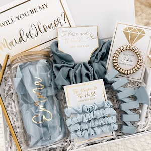 May include: A gift box filled with items for a maid of honor. The box includes a clear glass water bottle with a gold lid, a blue scrunchie, a blue hair claw, a gold hair tie, and a card that says "To have and to hold... your hair back".