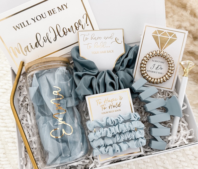 May include: A gift box filled with items for a maid of honor. The box includes a clear glass water bottle with a gold lid, a blue scrunchie, a blue hair claw, a gold hair tie, and a card that says "To have and to hold... your hair back".