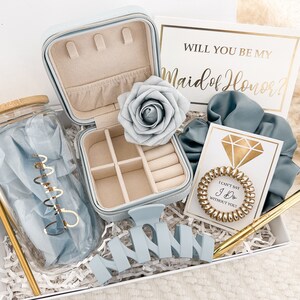 May include: A blue and gold bridesmaid gift box with a jewelry box, a glass jar with a blue ribbon, a blue scrunchie, a gold pen with a diamond-shaped top, a gold ring, a blue flower, and a card that says "Will you be my Maid of Honor?"