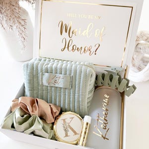 May include: A white gift box with gold detailing, containing a sage green cosmetic bag, scrunchies, a compact mirror, a lip balm, and a tumbler. The box lid reads "Will You Be My Maid of Honor?" in gold script. The cosmetic bag is embroidered with the name "Kat".