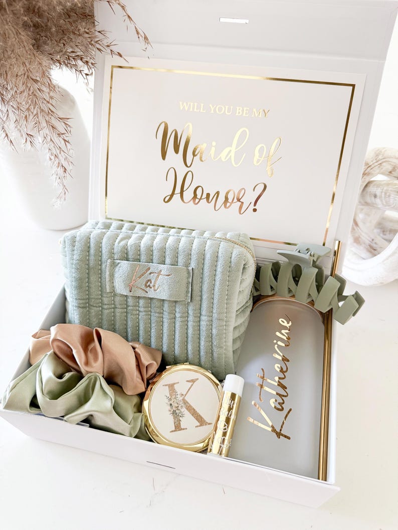 May include: A white gift box with gold detailing, containing a sage green cosmetic bag, scrunchies, a compact mirror, a lip balm, and a tumbler. The box lid reads "Will You Be My Maid of Honor?" in gold script. The cosmetic bag is embroidered with the name "Kat".