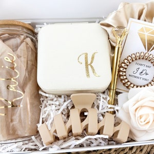 May include: A gift box filled with items for a bridal shower or wedding. The box includes a glass jar with a gold inscription that says "Katie", a small white jewelry box with a gold "K" on the lid, a gold pen, a gold hair tie, a white card with a gold diamond and the text "I can't say I Do without you", a beige hair clip, and a white rose.