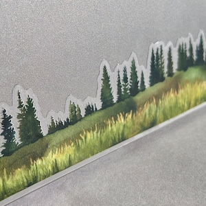 May include: A sticker of a watercolor landscape with a row of green trees and a field of green and yellow grass on a silver laptop.