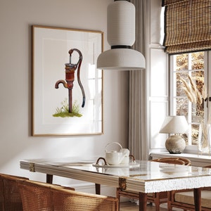 May include: A watercolor painting of a rusty hand pump with a wooden handle, set against a white background. The painting is framed in a light wood frame and hangs on a white wall above a dining table.