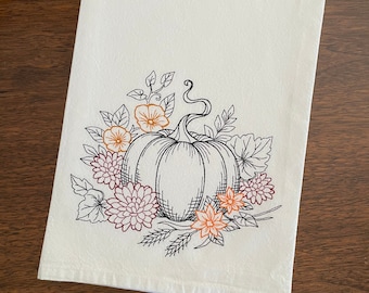 May include: A white kitchen towel with an embroidered design of a pumpkin surrounded by flowers and leaves. The pumpkin is outlined in black and the flowers are in shades of orange, yellow, and red.