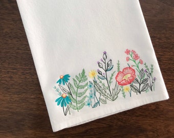 May include: White kitchen towel with embroidered floral design. The design features a variety of flowers in shades of blue, pink, yellow, and green.