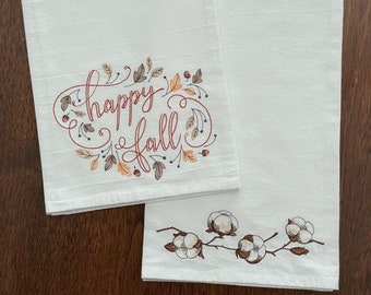 May include: Two white kitchen towels with embroidered designs. One towel features the text "happy fall" surrounded by brown and orange leaves. The other towel features a branch with white cotton bolls.
