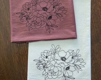May include: Two kitchen towels with a floral design embroidered on them. The top towel is a dusty rose color and the bottom towel is white.