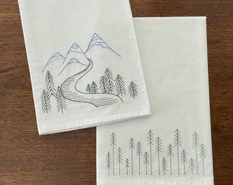 May include: Two white tea towels with embroidered designs. One towel features a mountain range with a winding road and trees. The other towel features a row of pine trees.
