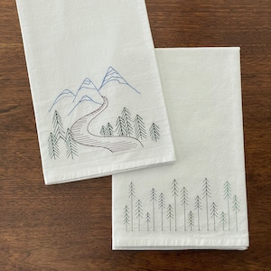 May include: Two white tea towels with embroidered designs. One towel features a mountain range with a winding road and trees. The other towel features a row of pine trees.