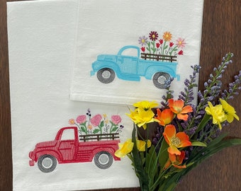 May include: Two embroidered kitchen towels with a red and blue pickup truck carrying flowers. The trucks are on a white background.