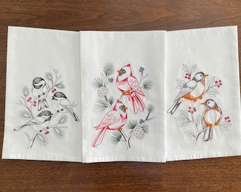 May include: Set of three white kitchen towels with embroidered winter bird designs. One towel features three black-capped chickadees on a branch with red berries. Another towel features two red cardinals on a branch with pine cones and berries. The third towel features two robins on a branch with holly berries.