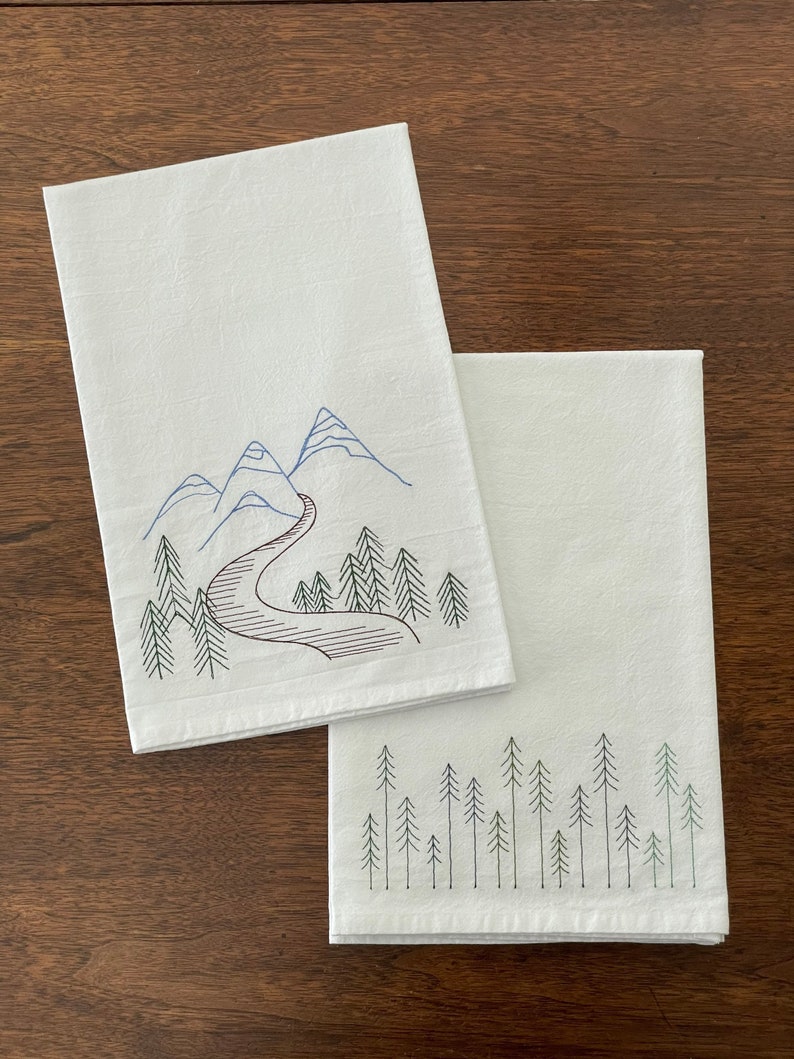 May include: Two white tea towels with embroidered designs. One towel features a mountain range with a winding road and trees. The other towel features a row of pine trees.