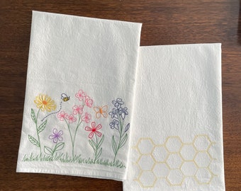 May include: Two white kitchen towels with embroidered designs. One towel features a bee and colorful flowers, while the other has a yellow honeycomb pattern.
