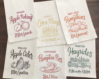 May include: Six white tea towels with fall-themed designs and prices. The designs include apple picking, corn maze, pumpkins, mulled apple cider, pumpkin pies, and hayrides.