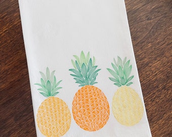 May include: A white kitchen towel with three embroidered pineapples. The pineapples are yellow, orange, and green.