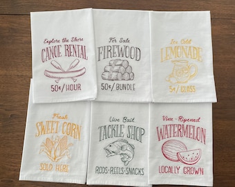 May include: Six white tea towels with embroidered designs and text. The designs include a canoe, firewood, a pitcher of lemonade, corn, a fish, and a watermelon. The text includes "Explore the Shore Canoe Rental 50¢/hour", "For Sale Firewood 50¢/bundle", "Ice Cold Lemonade 5¢/glass", "Fresh Sweet Corn Sold Here", "Live Bait Tackle Shop Rods-Reels-Snacks", and "Vine-Ripened Watermelon Locally Grown".