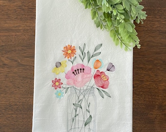 May include: A white kitchen towel with an embroidered design of a mason jar filled with colorful flowers. The flowers are pink, red, orange, yellow, and blue.