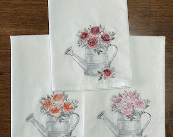 May include: Three white tea towels with embroidered images of watering cans filled with flowers. The watering cans are gray and the flowers are red, orange, and pink.