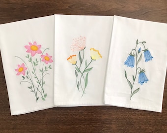 May include: Three white tea towels with embroidered floral designs. The first towel features pink flowers, the second towel features yellow and orange flowers, and the third towel features blue bell flowers.