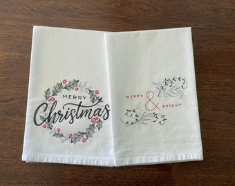 Puede incluir: Dos toallas de cocina blancas con diseños navideños bordados. Una toalla presenta una corona con el texto "Merry Christmas" y la otra toalla presenta un diseño floral con el texto "Merry & Bright".
