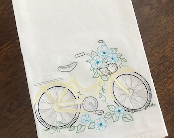 May include: White kitchen towel with an embroidered design of a yellow bicycle with a basket of blue flowers. The bicycle has black wheels and green vines.