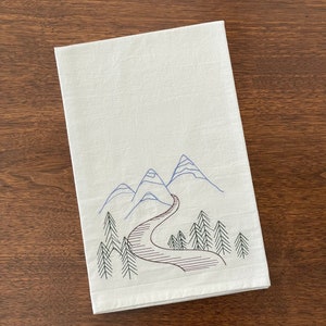 May include: White kitchen towel with an embroidered design of blue mountains and a winding brown road leading through a forest of green trees.
