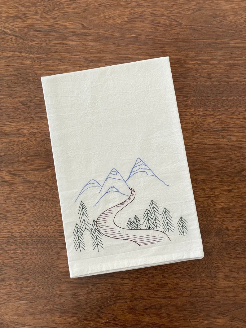 May include: White kitchen towel with an embroidered design of blue mountains and a winding brown road leading through a forest of green trees.