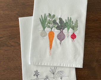May include: Two white kitchen towels with embroidered images of vegetables. The top towel features a row of five vegetables in color, including a carrot, onion, and beet. The bottom towel features a row of four vegetables in black and white, including a carrot, onion, and beet.