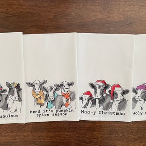 Peut inclure: Quatre torchons de cuisine blancs avec des illustrations de vaches. Chaque torchon présente un design de vache différent avec le texte : "Udderly fabulous", "Herd it's pumpkin spice season", "Moo-y Christmas" et "Holy cow, it's cold."