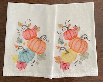 May include: Two white kitchen towels with embroidered fall-themed designs. Each towel features a cluster of three pumpkins in shades of orange, yellow, and blue, surrounded by leaves in shades of red, green, and brown.
