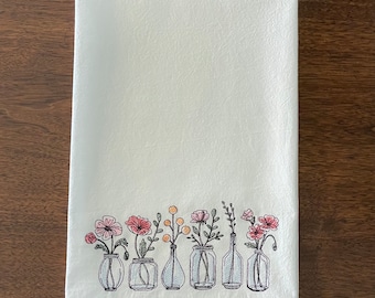 May include: A white kitchen towel featuring an embroidered design along the bottom edge. The embroidery depicts six clear glass vases of varying shapes, each containing a different botanical element, including pink flowers and yellow buds.