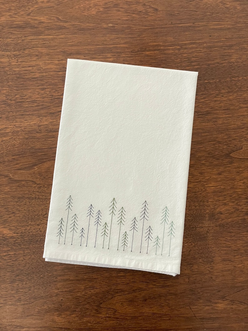 May include: White kitchen towel with a green embroidered design of a row of pine trees.