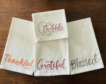 May include: Set of four white kitchen towels with embroidered Thanksgiving-themed designs. The towels feature the words "Gobble", "Thankful", "Grateful", and "Blessed" in different colors and fonts.
