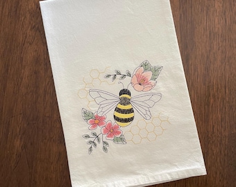May include: White kitchen towel with an embroidered design featuring a bee, pink flowers, and a honeycomb pattern. The bee has black and yellow stripes. The flowers have pink petals and green leaves.