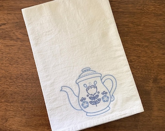 May include: White tea towel with a blue embroidered teapot design. The teapot features a floral motif and is centered on the towel. The towel is folded and lies on a wooden surface.