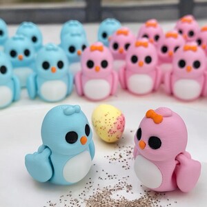 May include: A collection of small, hand-painted ceramic bird figurines. The birds are in shades of blue and pink, with white bellies and black eyes. Some of the birds have orange beaks and orange bows. There is a speckled egg in the center of the image.