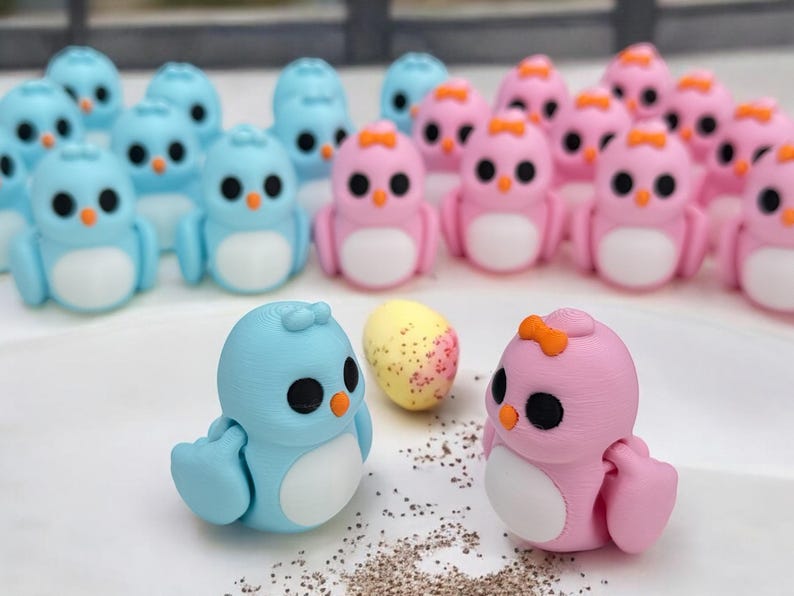 May include: A collection of small, hand-painted ceramic bird figurines. The birds are in shades of blue and pink, with white bellies and black eyes. Some of the birds have orange beaks and orange bows. There is a speckled egg in the center of the image.