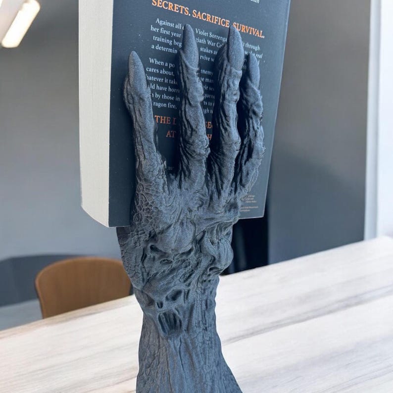 May include: A dark gray, textured hand-shaped bookend holding a book. The bookend has a detailed, gnarled appearance, with visible veins and a rough surface. The book's title is partially visible, with the words "SECRETS. SACRIFICE SURVIVAL."
