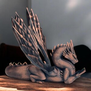 May include: A gray dragon figurine with wings outstretched, reading a book. The dragon is sitting on a wooden surface.