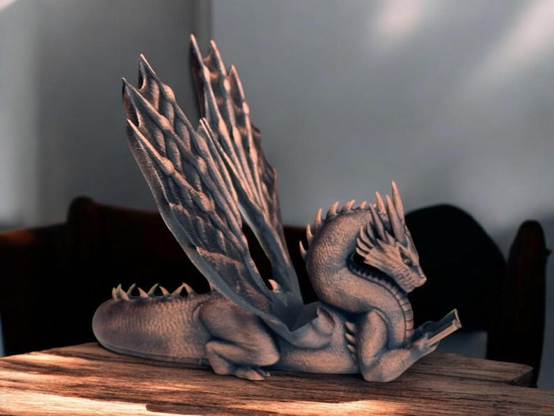 May include: A gray dragon figurine with wings outstretched, reading a book. The dragon is sitting on a wooden surface.