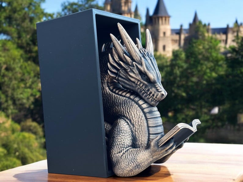 May include: A silver dragon bookend with a detailed, textured design. The dragon is holding a book in its claws and is positioned in a reading pose. The bookend is made of a durable material and is perfect for holding books upright on a shelf or desk.