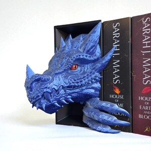 May include: A blue dragon head bookend with red eyes and sharp teeth. The dragon's head and claw extend from a black bookend, positioned between books with the author's name "SARAH J. MAAS" on the spine. The bookend adds a fantasy theme.