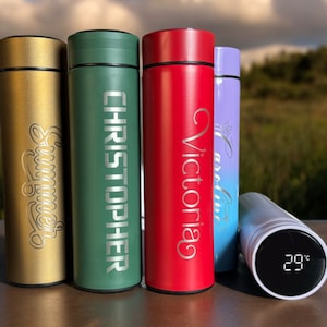 Five colorful thermos bottles with personalised names: A white bottle with a digital display reading "29°C". Blurred background.