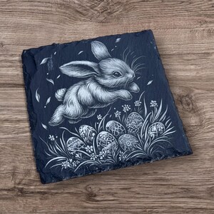 Engraved slate coaster featuring a detailed illustration of a bunny resting on a decorative Easter egg, surrounded by grass and leaves. The coaster has a natural, rough-edged finish and is set on a wooden background