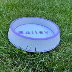 May include: A white and purple pet bowl with the name "Bailey" printed on the inside. The bowl is sitting on a bed of green grass.