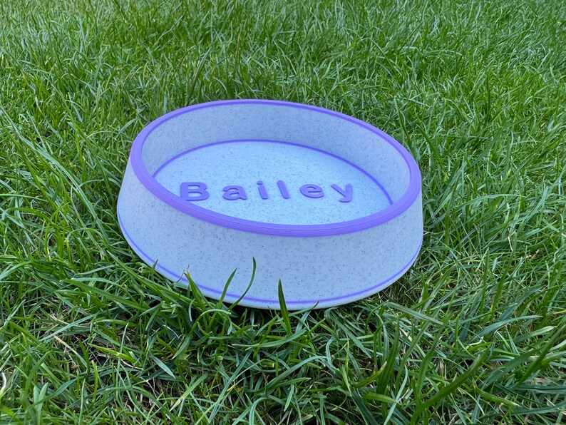 May include: A white and purple pet bowl with the name "Bailey" printed on the inside. The bowl is sitting on a bed of green grass.